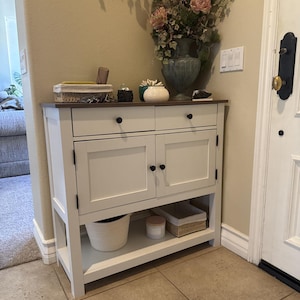 May include: A white wooden console table with a dark brown top, featuring two upper drawers with black knobs and two lower cabinet doors with black knobs. The table has an open bottom shelf holding a white woven basket and a small rectangular basket.