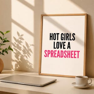 Könnte beinhalten: Ein gerahmter Druck mit dem Text "HOT GIRLS LOVE A SPREADSHEET" in Schwarz und Pink. Ein Laptop, eine Kaffeetasse und eine Vase mit Grünzeug stehen auf einem weißen Schreibtisch, der von natürlichem Licht beleuchtet wird.