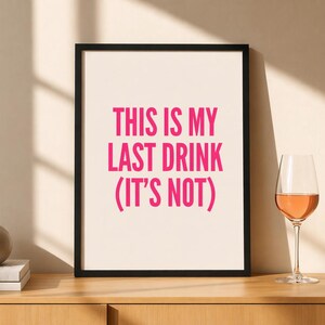 Könnte beinhalten: Gerahmter Druck mit dem Text "THIS IS MY LAST DRINK (IT'S NOT)" in leuchtendem Pink. Ein Weinglas mit Roséwein steht auf einer Holzoberfläche, neben einer kleinen grauen Vase und einem Stapel Bücher.