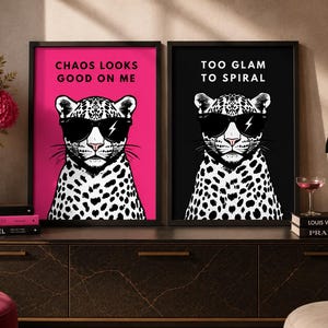Könnte beinhalten: Zwei gerahmte Kunstdrucke mit Leoparden-Illustrationen, die eine Sonnenbrille tragen. Einer hat einen rosa Hintergrund mit dem Text "CHAOS LOOKS GOOD ON ME". Der andere hat einen schwarzen Hintergrund mit dem Text "TOO GLAM TO SPIRAL".
