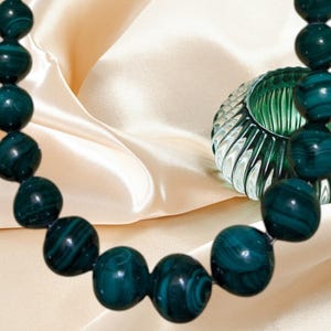 May include: A necklace composed of round, dark green beads featuring natural swirling patterns, arranged in a curved line. Beside the beads sits a small, translucent, ribbed green glass bowl with a rounded shape and vertical ridges.