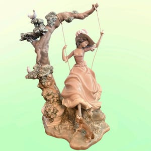 May include: A detailed porcelain figurine of a woman on a swing, wearing a pink dress and hat. The swing is attached to a tree with two doves perched on its branches. A small dog stands near the woman. The figurine is in shades of pink, brown, and green.