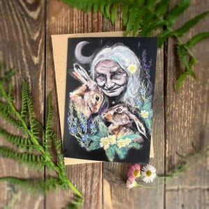 May include: A rectangular greeting card featuring a black background with a detailed illustration of an elderly person with long white hair, two brown rabbits, a crescent moon, and colorful floral accents in shades of blue, yellow, and green.