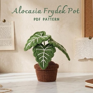 May include: A crocheted Alocasia Frydek plant in a brown pot. The plant has green leaves with white veins. The text "Alocasia Frydek Pot PDF Pattern" is above the plant. The background includes a corkboard, artwork, and a notebook.