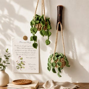 May include: Two hanging crochet planters with green trailing vines and heart-shaped leaves. The planters are made of tan yarn and suspended by braided cords. A white paper print with the text Forrest Gump and botanical illustrations is nearby.