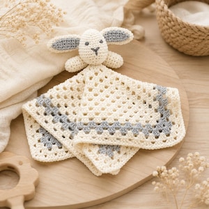 Puede incluir: Una manta de seguridad de ganchillo con una base cuadrada de granny square en color crema y un borde gris. En el centro hay una cabeza de conejo de ganchillo rellena con orejas largas y ojos y nariz bordados en gris oscuro.