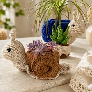 May include: Two crocheted snail-shaped planters. The front snail has a beige head and a brown shell containing purple succulents. The back snail has a beige head and a blue shell containing a green crocheted plant. Both have black eyes.