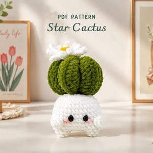 Puede incluir: Un cactus de ganchillo hecho a mano con una base blanca, un cuerpo verde y una flor blanca. El cactus tiene ojos negros y mejillas rosas. El texto "PDF PATTERN Star Cactus" está en la parte superior. El cactus está sobre una superficie blanca.