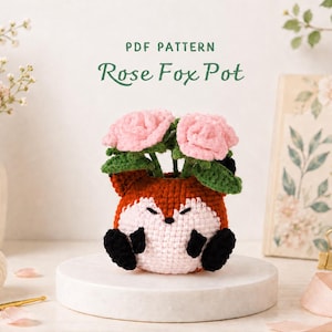 May include: A crochet fox-shaped pot featuring a rust-orange and light pink body with black paws and ears. The pot contains two crocheted light pink roses with green leaves. The text PDF PATTERN Rose Fox Pot is displayed above the object.