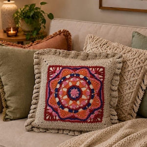 Pode incluir: Uma almofada quadrada de crochê com um design floral colorido em tons de vermelho, laranja, rosa e azul. A almofada tem uma borda bege e está colocada em um sofá bege. Outras almofadas em várias texturas e cores também são visíveis.