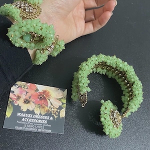 May include: Two beaded bracelets in a light green hue, with gold-colored accents and decorative embellishments. The bracelets are adorned with small, round beads. A business card with floral designs and the text "WAKUKI DRESSES & ACCESSORIES" is also visible.