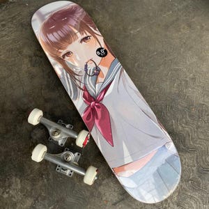 Może przedstawiać: Deska do skateboardu z ilustracją w stylu mangi przedstawiającą dziewczynę w mundurku szkolnym. Deska ma 20,3 cm szerokości. Obok deski znajdują się dwa srebrne trucki do skateboardu z białymi kółkami.