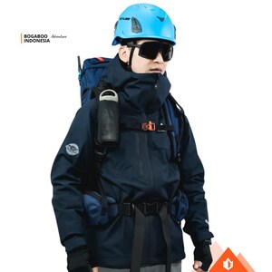 May include: A navy blue outdoor jacket with a matching backpack, water bottle, and a blue helmet. The jacket has a high collar and multiple pockets. The image includes the text "BOGABOO Adventure INDONESIA". The person is wearing black sunglasses.