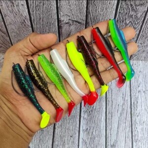 May include: A collection of colorful fishing lures in various shades of green, black, white, red, and blue. Each lure has a hook and a tail, designed to mimic small fish. The lures are held in a hand against a wooden background.