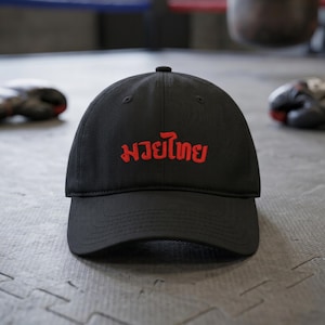 Peut inclure: Casquette de baseball noire avec le texte thaïlandais rouge "Muay Thai" brodé sur le devant. La casquette a une visière incurvée et est faite d'un matériau résistant. Des gants de boxe et un sol de gymnase sont visibles en arrière-plan.