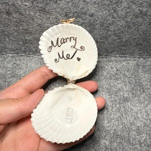 May include: A white seashell ring box with a hinged metal clasp. The interior of the top shell features the handwritten black text "Marry Me" with a small heart symbol. The bottom shell contains a clear plastic ring holder centered inside.