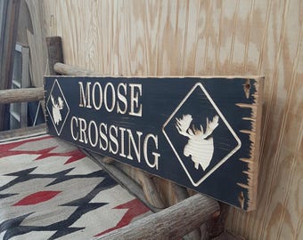 May include: A black wooden sign with the words "MOOSE CROSSING" in white lettering. The sign has a diamond-shaped warning sign with a moose silhouette in the center.
