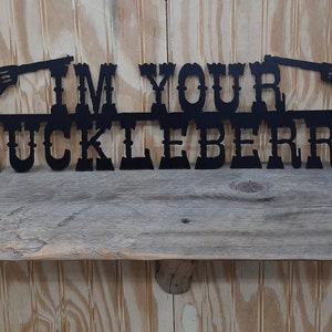May include: Black metal wall decor with the text "I'm Your Huckleberry" and two pistols on either side.