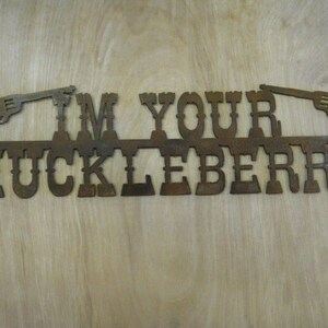 May include: Rustic metal sign with the text "I'm Your Huckleberry" and two stylized pistols on either side.