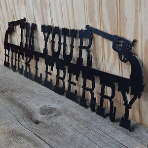 May include: Black metal wall decor with the text "I'm Your Huckleberry" and two stylized pistols.