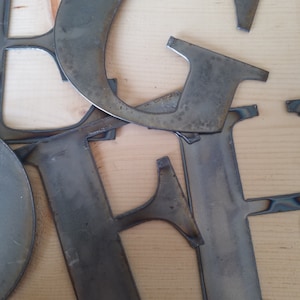 May include: Metal letters in the shape of the letters E, G, and F. The letters are made of a dark gray metal and have a rough, unfinished texture.