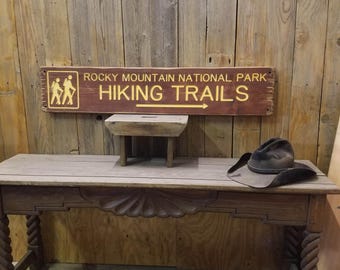 May include: A rustic wooden sign with the text "Rocky Mountain National Park Hiking Trails" in yellow lettering. The sign is mounted on a wooden wall and features a yellow silhouette of two hikers walking on a trail.