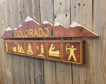 May include: Wooden sign with the word "COLORADO" in yellow letters on a brown background. The sign has a mountain range silhouette with snow-capped peaks. Below the sign is a row of six icons depicting outdoor activities: fishing, camping, campfire, hiking, boating, and archery.