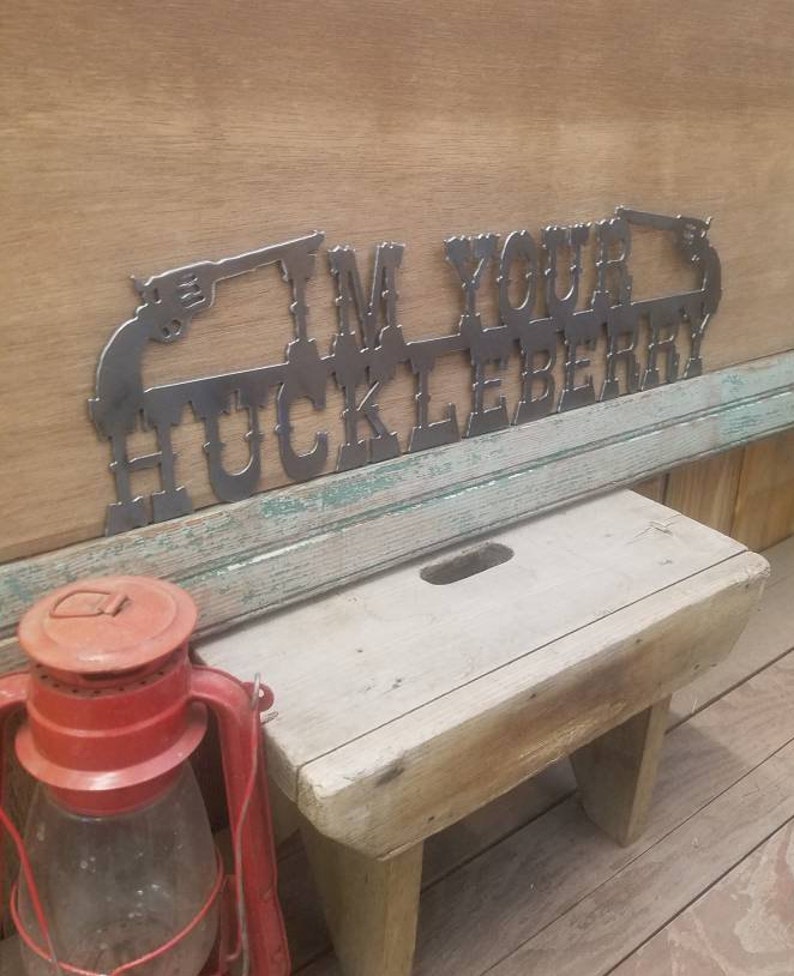 May include: Metal wall art with the text "I'm Your Huckleberry" and two stylized pistols on either side. The art is mounted on a wooden wall.