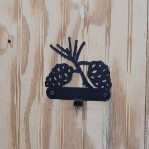 May include: Black metal pine cone hook with a single hook on the bottom. The hook is mounted on a wooden surface.
