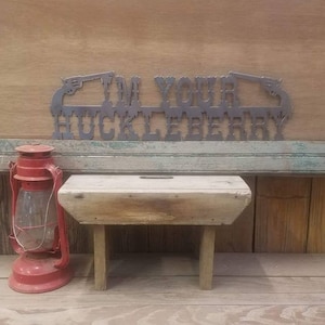 May include: A metal sign with the text "I'm Your Huckleberry" in a western style font. The sign is decorated with two pistols on either side of the text. The sign is hanging on a wooden wall.