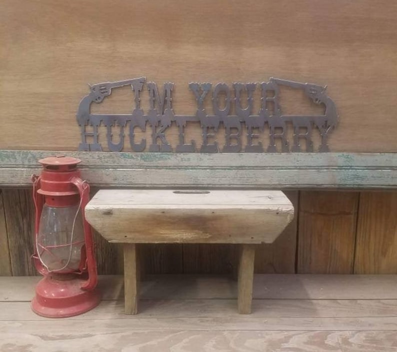 May include: A metal sign with the text "I'm Your Huckleberry" in a western style font. The sign is decorated with two pistols on either side of the text. The sign is hanging on a wooden wall.