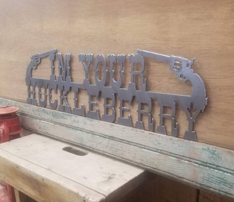 May include: Metal wall art with the text "I'm Your Huckleberry" and two stylized pistols. The art is painted in a silver color.