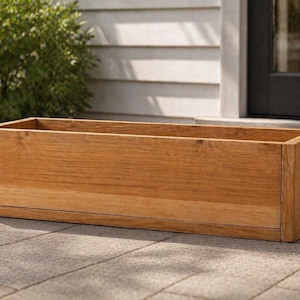 May include: A rectangular wooden planter box with a natural wood grain finish. The box is empty and sits on a stone patio. The planter is designed with clean lines and a simple, modern aesthetic, suitable for outdoor use.