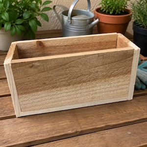 Op de afbeelding: Een rechthoekige houten plantenbak gemaakt van lichtbruine natuurlijke houten planken met zichtbare nerven. De bak heeft een eenvoudig ontwerp met een open bovenkant, versterkte verticale hoekranden en een platte bodem met een ruwe afwerking.