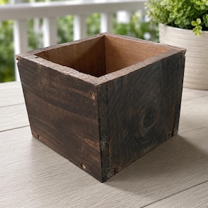 May include: A square, dark brown wooden box with visible wood grain. The box has a rustic appearance and is empty. The box is sitting on a light-coloured surface, with a white railing and greenery in the background.