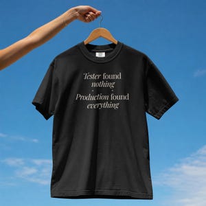 Könnte beinhalten: Ein schwarzes Kurzarm-T-Shirt mit weißem Text: Tester found nothing, Production found everything. Das Shirt hängt an einem Holzkleiderbügel vor einem klaren blauen Himmel und zeigt einen Rundhalsausschnitt sowie eine klassische Passform.
