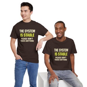 May include: A dark brown short-sleeved t-shirt featuring the text THE SYSTEM IS STABLE in cream and yellow, followed by the text PLEASE DON'T TOUCH ANYTHING in cream. The shirt has a crew neckline and a standard fit, displayed on two individuals.
