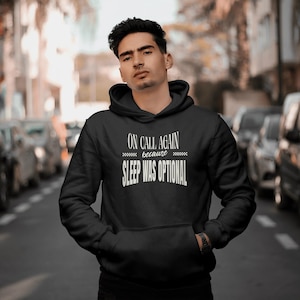 May include: A black hooded sweatshirt featuring white text on the chest that reads ON CALL AGAIN because SLEEP WAS OPTIONAL. The hoodie includes a front kangaroo pocket, long sleeves with ribbed cuffs, and a drawstring hood in a solid black fabric.