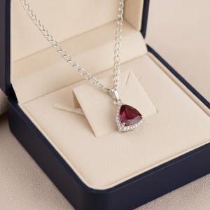 May include: A silver necklace with a triangular, deep red pendant. The pendant is surrounded by small, clear stones and is displayed in a blue jewelry box with a cream-colored interior.