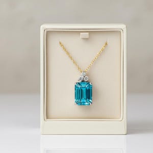 May include: A gold chain necklace with a large, rectangular, bright blue gemstone pendant. The pendant is set in a silver-colored metal and has three small, clear stones above it. The necklace is displayed in a cream-colored jewelry box.
