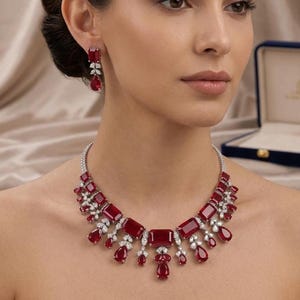 May include: A necklace and matching earrings set featuring deep red rectangular and pear-shaped gemstones accented with small clear stones. The necklace has a central rectangular stone with cascading red drops, while the earrings feature a single rectangular stone above a pear-shaped drop.