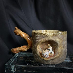 May include: A unique ceramic mug with a tree-like design and a branch handle. The mug features a small, detailed figurine of a calico cat nestled inside a hollowed-out section. The mug is brown and beige with a glossy finish.