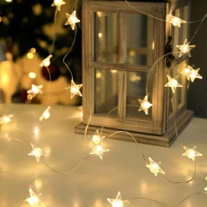 Può includere: Una stringa di luci a forma di stella bianco caldo. Le luci sono infilate su un filo sottile e trasparente e sono sparse su una superficie bianca. Una piccola lanterna decorativa in legno è sullo sfondo.