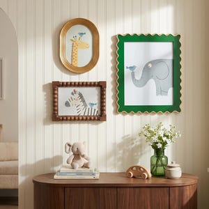 May include: A collection of framed animal art, including a giraffe, zebra, and elephant, each with a small blue bird. The frames are gold, green, and brown. A plush elephant toy and wooden car sit on a wooden cabinet.