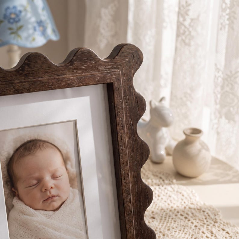 May include: A dark brown wooden picture frame with a scalloped edge design. The frame contains a white mat and a photograph of a sleeping infant wrapped in a white blanket. The wood grain is visible on the frame's surface, highlighting its texture.
