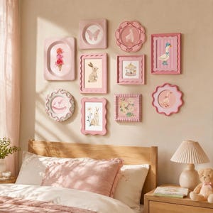 Op de afbeelding: Een verzameling roze ingelijste wandkunst met verschillende illustraties, waaronder een vlinder, konijn, giraf en bloemen. De lijsten hebben verschillende vormen, waardoor een speelse opstelling ontstaat. De kunst hangt aan een beige muur boven een bed met roze beddengoed.
