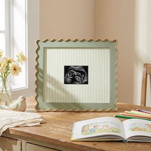 Puede incluir: Un marco de fotos rectangular con borde festoneado y acabado en verde salvia, que presenta un borde interior dorado. El marco contiene un paspart&uacute; de color claro con rayas verticales y una imagen de ecograf&iacute;a en blanco y negro.