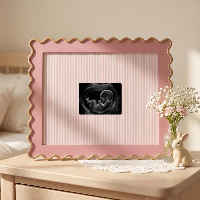 Puede incluir: Un marco de fotos rosa con bordes festoneados que contiene una ecograf&iacute;a en blanco y negro de un feto. El marco presenta un paspart&uacute; con rayas verticales de color rosa claro y blanco. Se exhibe junto a un peque&ntilde;o florero de cristal.