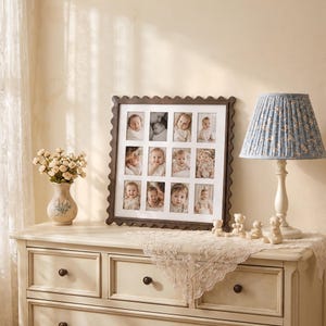 May include: A square dark wood picture frame with a scalloped edge containing a white mat with twelve rectangular openings displaying black and white photographs of infants. The frame is positioned on a light-colored wooden dresser surface.