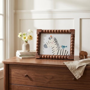 K&ouml;nnte beinhalten: Ein rechteckiger Bilderrahmen mit dunkelbraunem Holzfinish und einem dekorativen Perlenrand. Der Rahmen enth&auml;lt einen Druck eines stilisierten Zebras mit einem kleinen blauen Vogel auf dem R&uuml;cken vor einem schlichten wei&szlig;en Hintergrund.